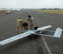Three soldiers work on the Shadow UAV in the middle of a runway, one soldier is holding a long a device with a long antenna and a laptop sits on the UAV. Two civilian vehicles are parked on the edge of the runway in the background. Low cloud and fog can b